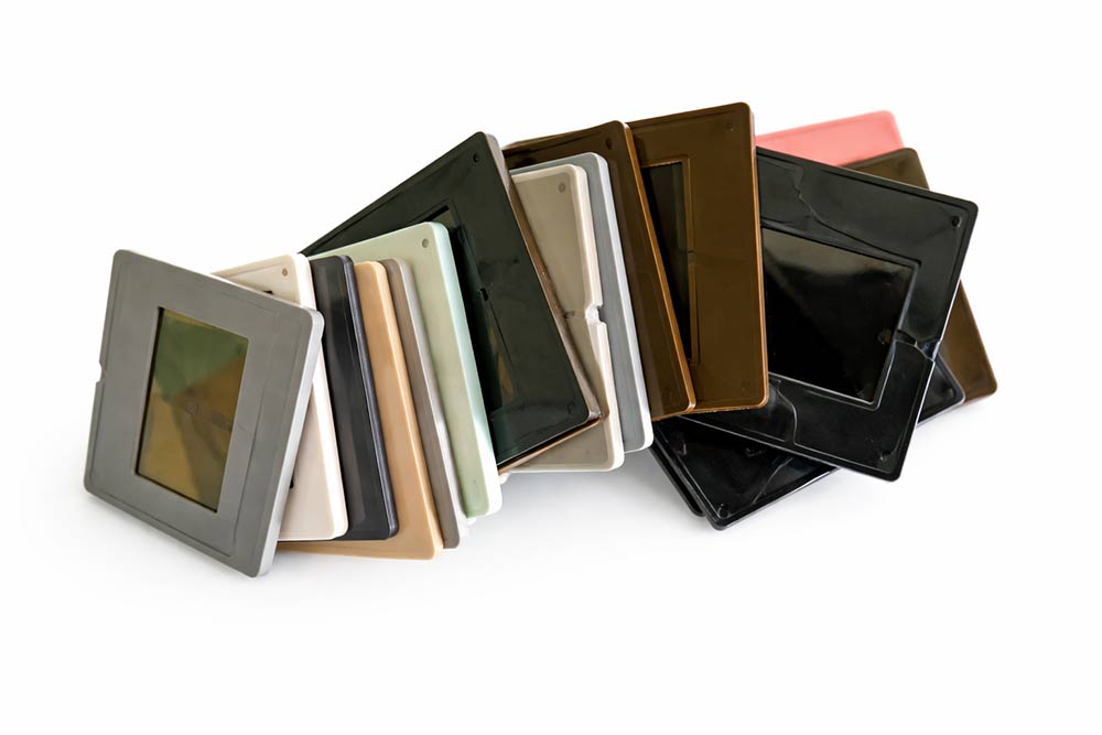 Оцифрування діапозитивів у Києві — точний колір та висока деталізація кожного слайда 5 Ряд фотографічних слайдів у пластикових рамках різних кольорів (сірий, коричневий, бежевий, чорний), викладених віялом на білому фоні.